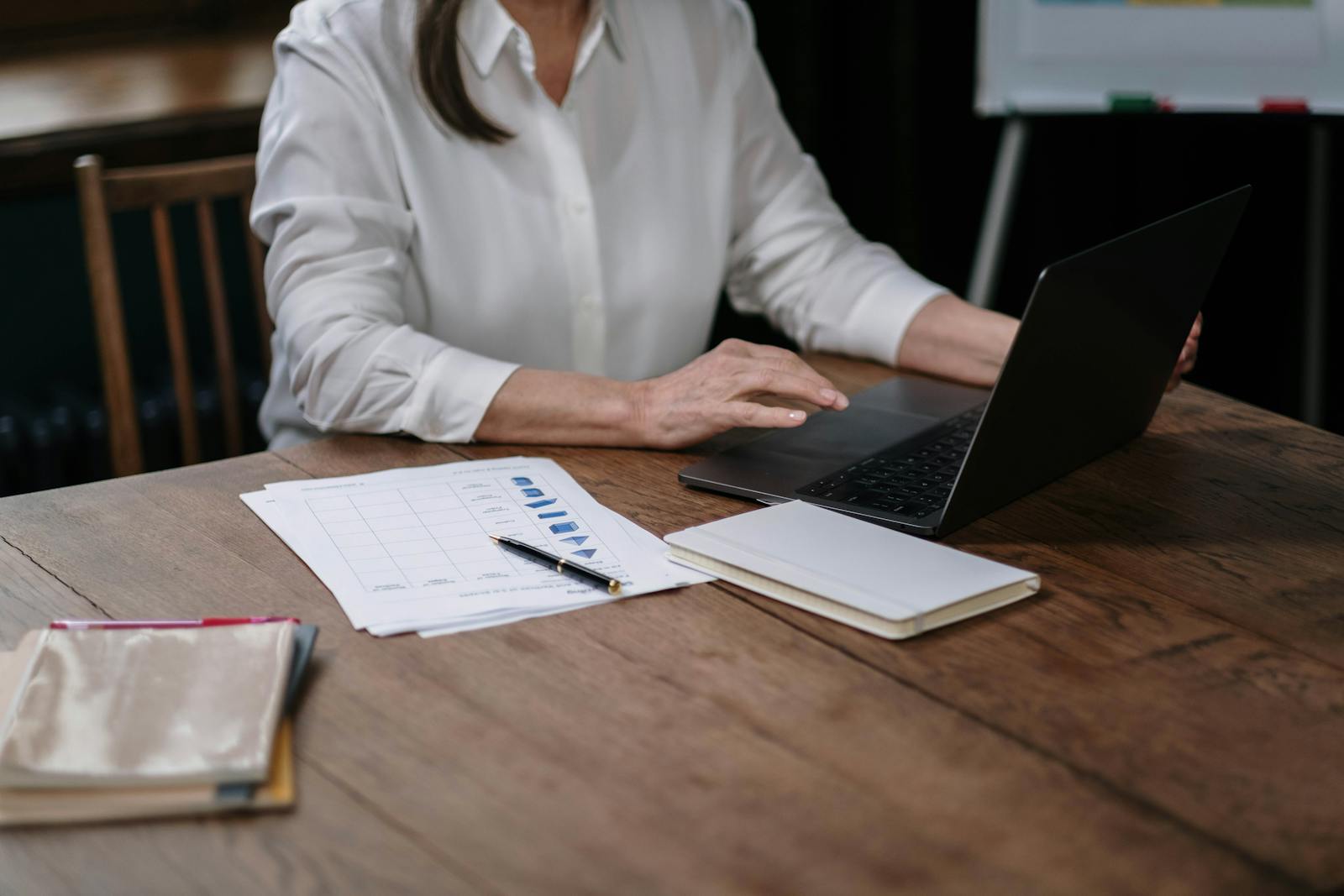 Businesswoman in white shirt working on laptop with papers at wooden desk.